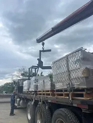 A crane lifts a pallet-moving attachment above a flatbed truck loaded with wrapped cargo, while a person stands nearby.