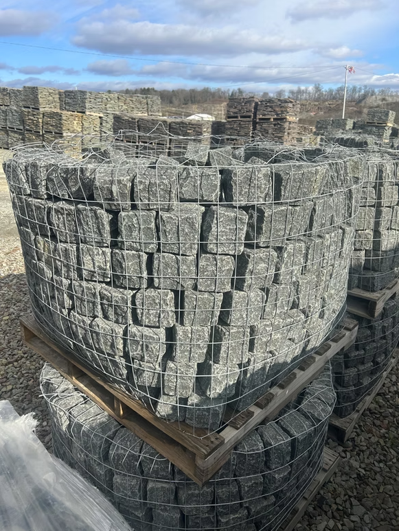 Gray granite cobblestones stacked in wire cages on wooden pallets in an outdoor stone yard.