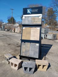 A crane lifts a pallet-moving attachment above a flatbed truck loaded with wrapped cargo, while a person stands nearby.