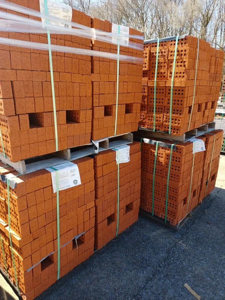 Stacked pallets of rectangular, red clay bricks with hole patterns, secured by plastic strapping outdoors.