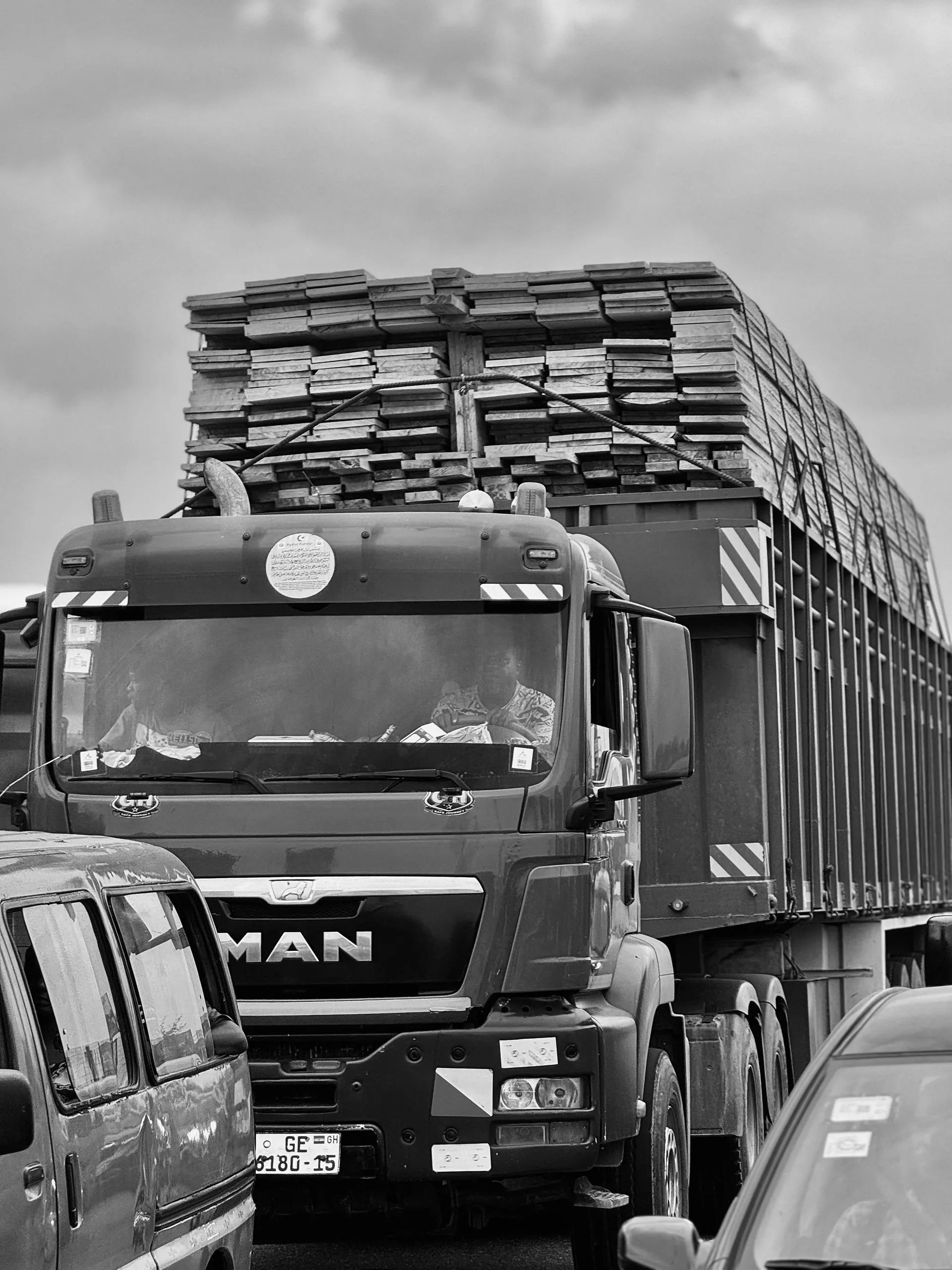A black and white photo of a MAN truck loaded with stacks of lumber driving on a road surrounded by other vehicles.