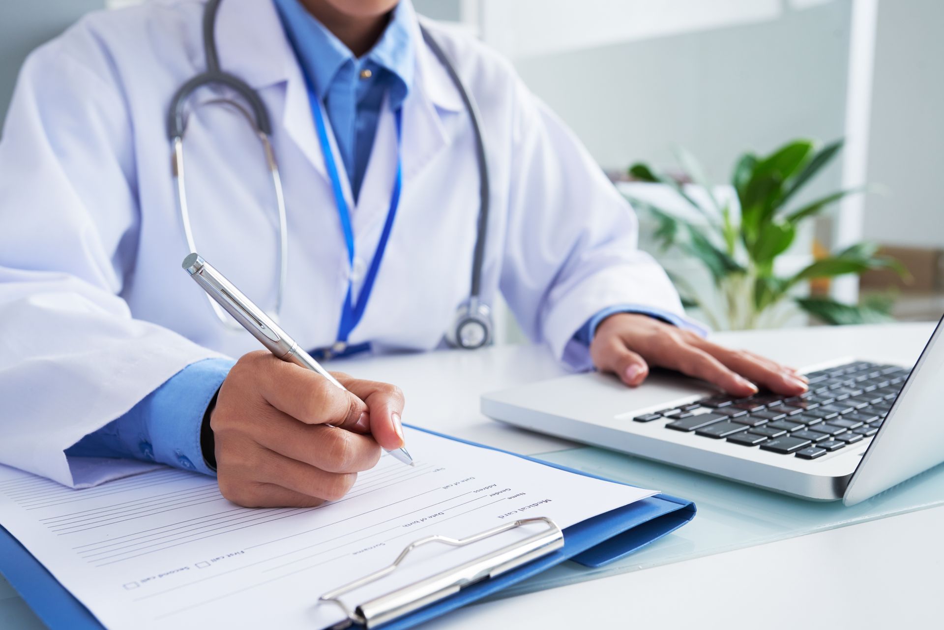 Um médico de jaleco branco escreve em uma prancheta enquanto usa um laptop em uma mesa.