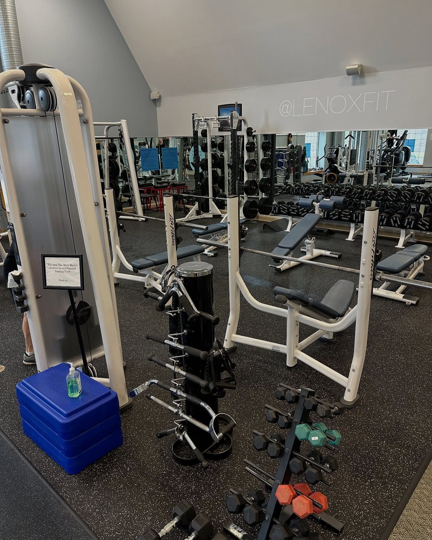 An indoor gym featuring weight benches, barbell racks, dumbbell towers, and a blue step platform on rubber flooring.