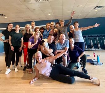 A diverse group of people posing and smiling in a fitness studio with exercise equipment in the background.