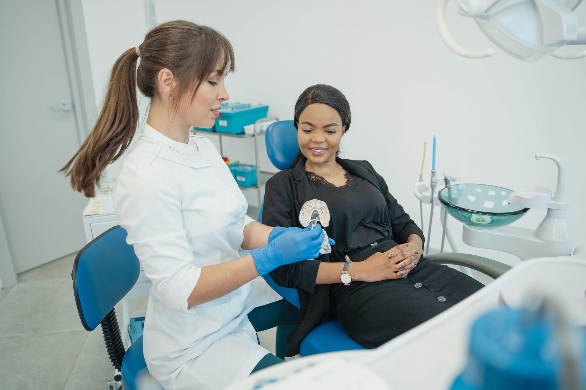 A dentist in blue gloves shows a patient a clear dental retainer in a bright office. The patient smiles.