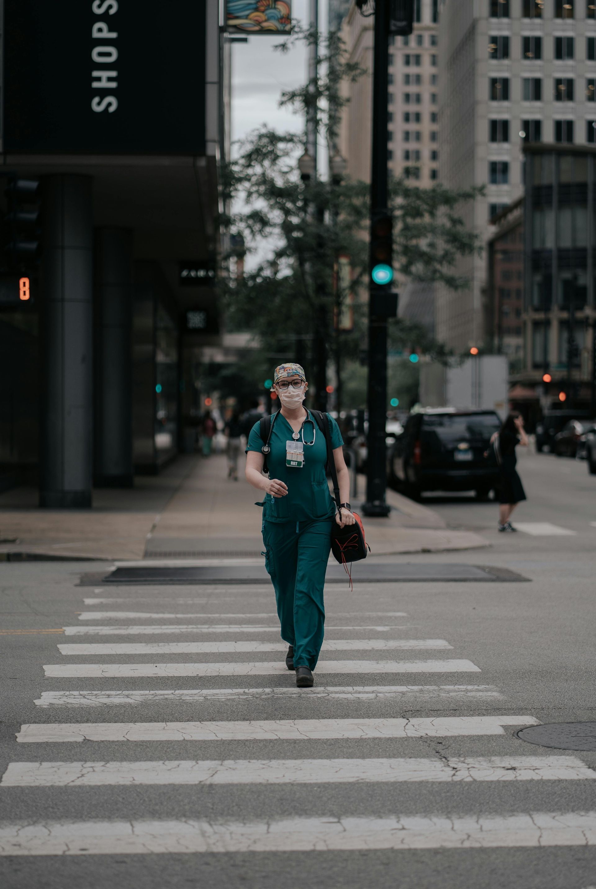 A healthcare worker in green scrubs crosses a city street, carrying a bag, with a facemask and glasses.