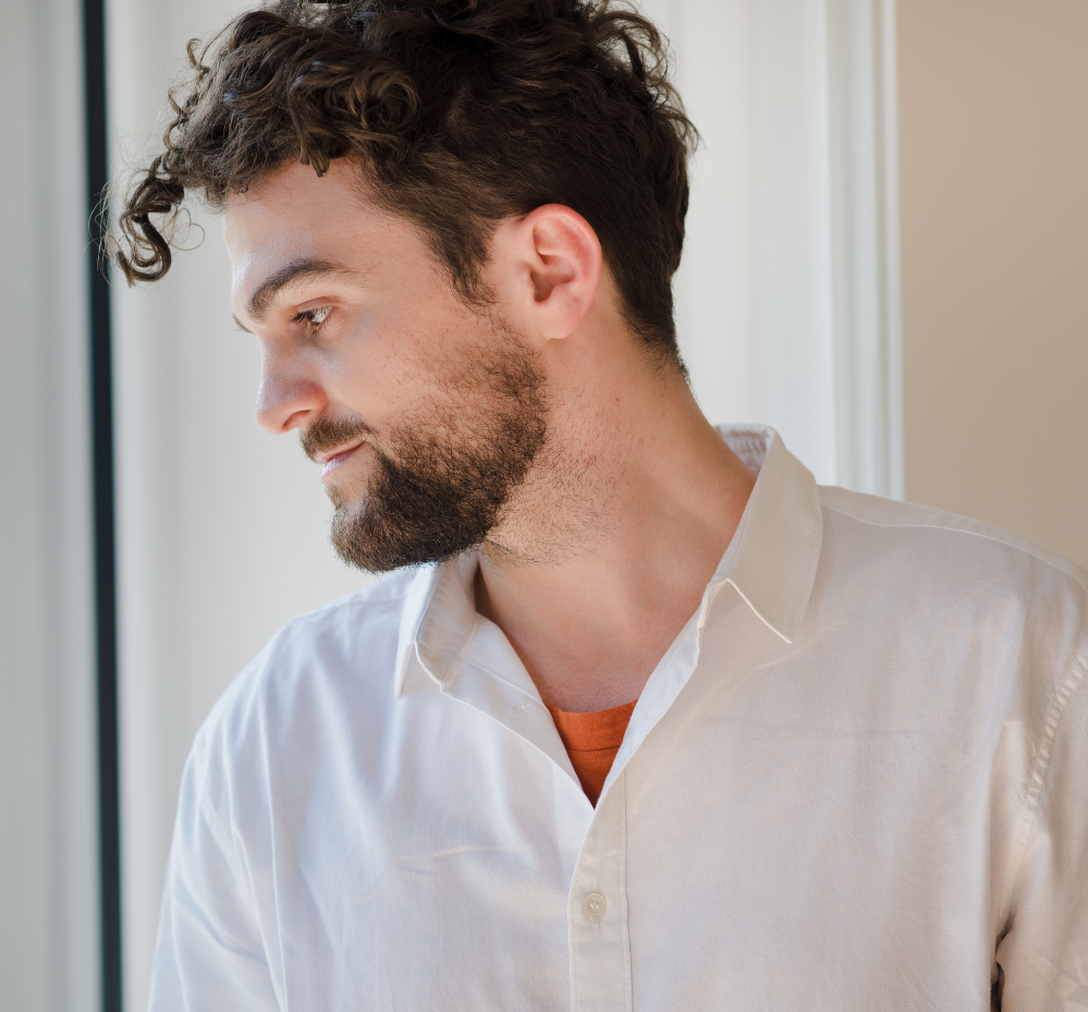 Man with curly hair and beard in a white shirt, looking out a window.
