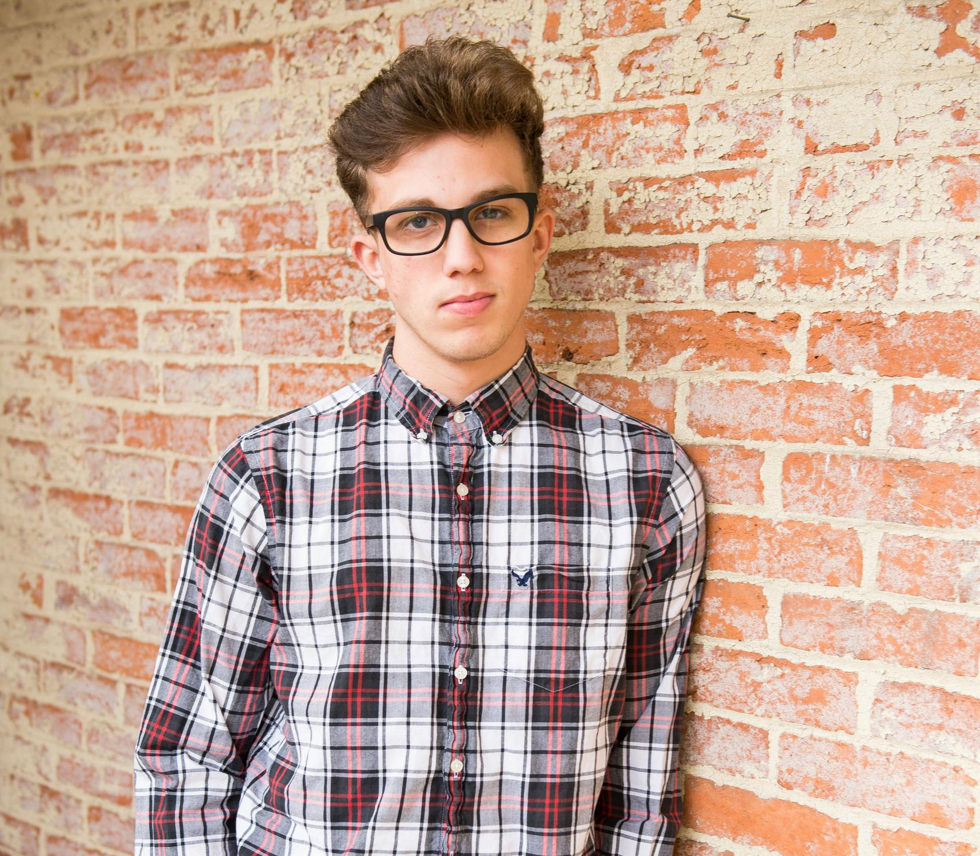 Young man in glasses and plaid shirt leaning against a brick wall, hands in pockets.