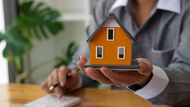 A real estate agent, wearing a gray suit, holds a small house model as they sit inside an office.