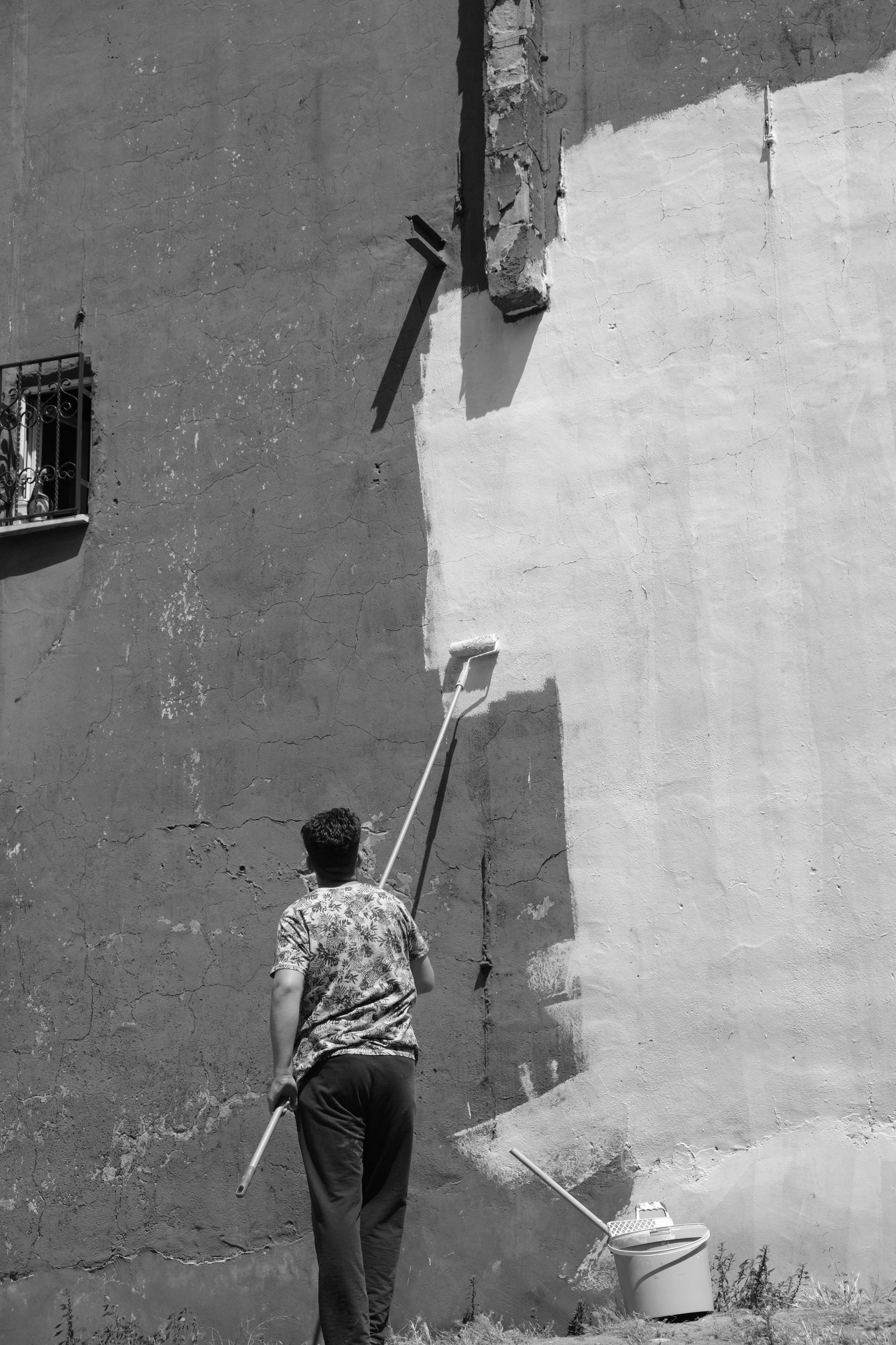 A person paints a wall white with a long-handled roller, with a paint bucket on the ground nearby.