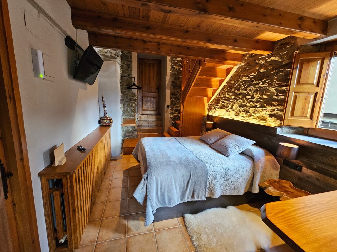 Dormitori acollidor amb bigues de fusta a la vista, paret de pedra, llit, petita catifa i escala. Cal Serni