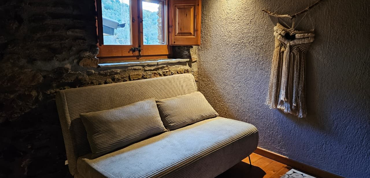 Un petit sofà es troba sota una finestra amb persianes de fusta. A prop hi ha una decoració de paret.