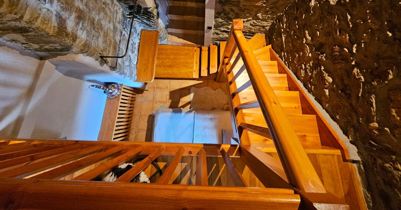 Escala de fusta en una habitació amb parets de pedra, il·luminada per una llum càlida. Cal Serni