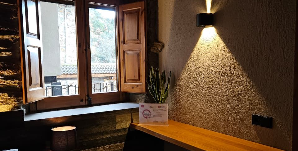 Racó interior amb finestra oberta, banc de fusta i aplic de paret modern, tons càlids. Cal Serni