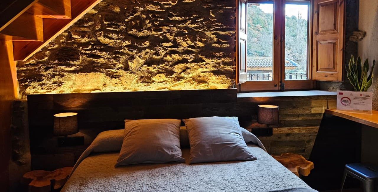 Dormitori amb paret de pedra vista, somier de fusta i finestra amb vistes a una muntanya. Cal Serni