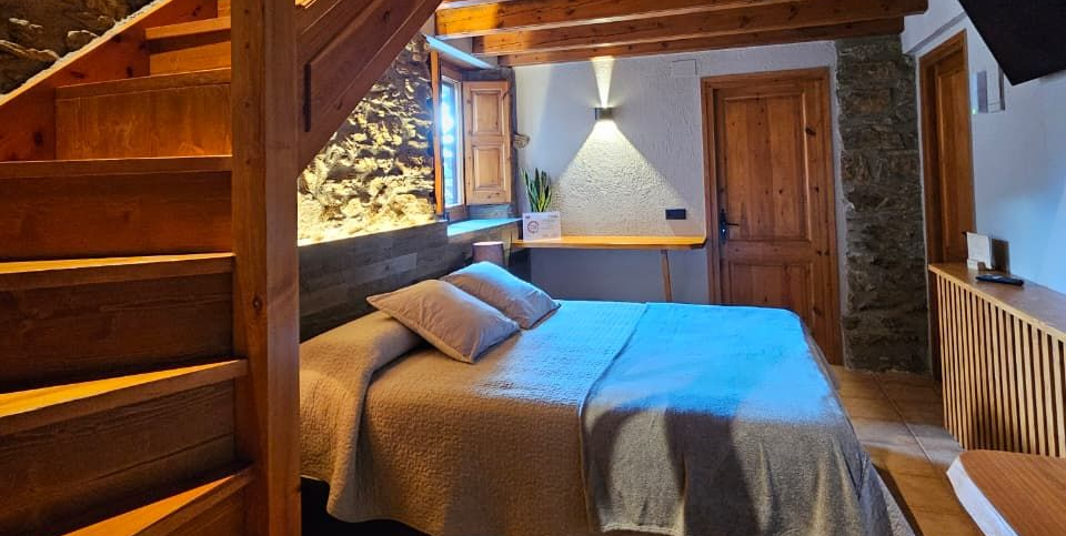 Dormitori amb escala de fusta, llit i paret de pedra, imatge interior. Cal Serni