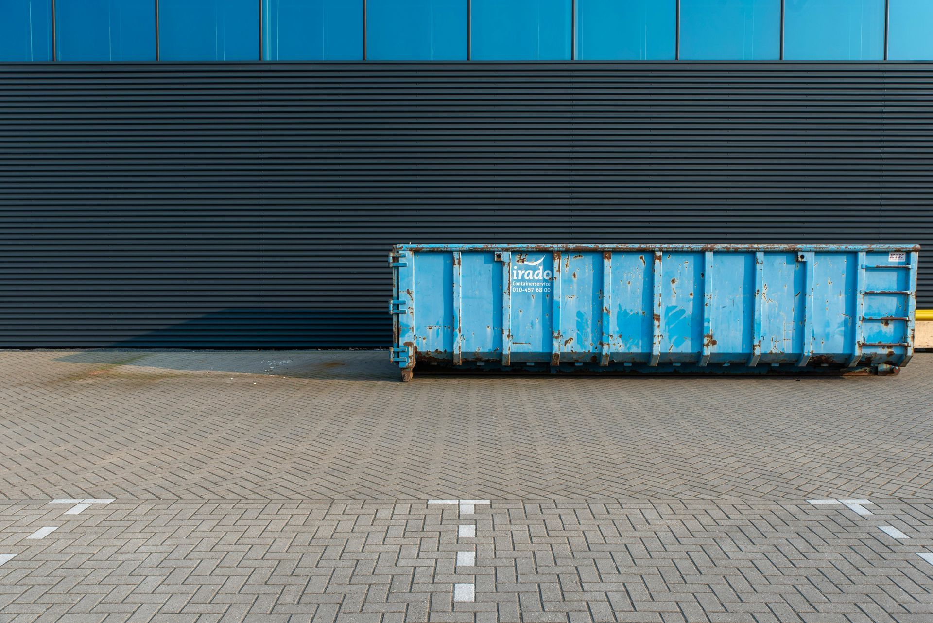 Blue dumpster against a dark, ribbed wall and blue glass windows in a paved parking area.