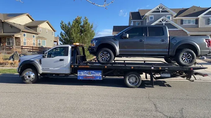 Silver tow truck carrying a gray Ford pickup truck on a residential street. Bright, sunny day.