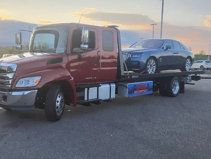 Red tow truck hauling a blue Rolls-Royce on a flatbed trailer in a parking lot. Evening sky.
