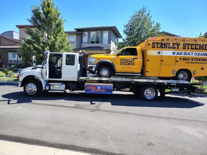 A tow truck hauling a yellow Stanley Steemer work truck on a residential street.