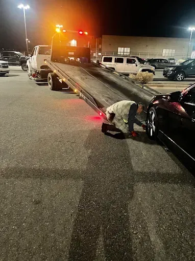 Tow truck loading a black sports car at night in a parking lot. A person is working at the car.