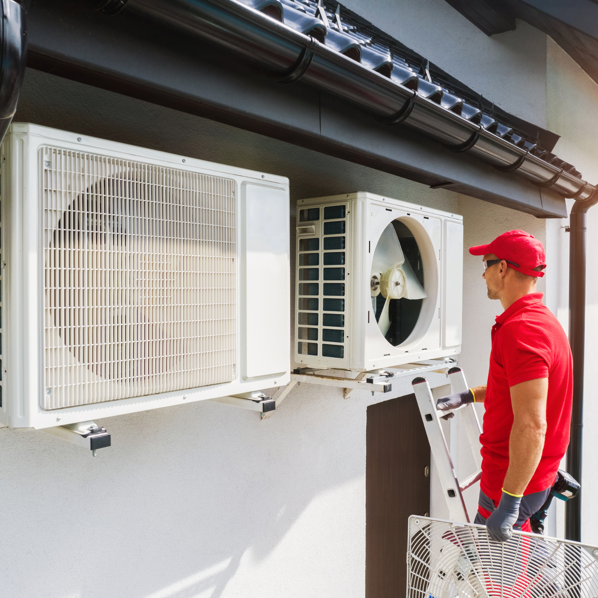 HVAC technician in red shirt and cap on ladder, servicing air conditioning units on a white wall.