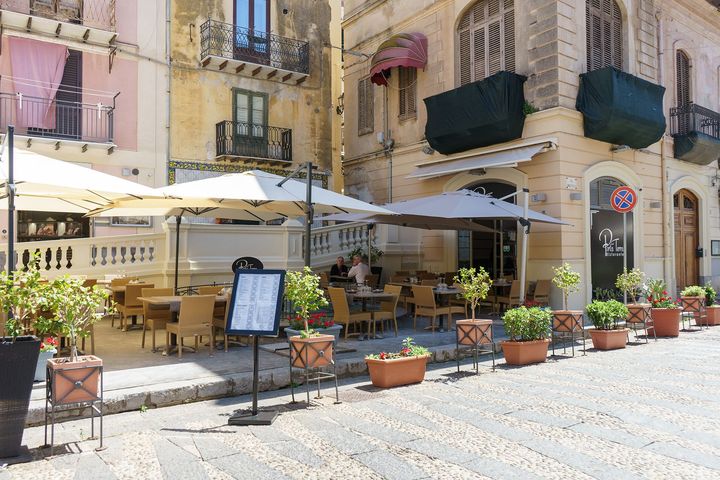 Caffè all'aperto con tavoli, ombrelloni, piante in vaso ed edifici in pietra in una piazza soleggiata