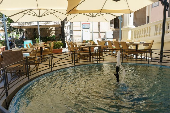 Dehor esterno del caffè con piccola fontana, tavoli, sedie e grandi ombrelloni.