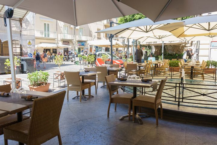 Terrazza esterna di un caffè con tavolini, sedie e grandi ombrelloni in legno, situata in una strada soleggiata.