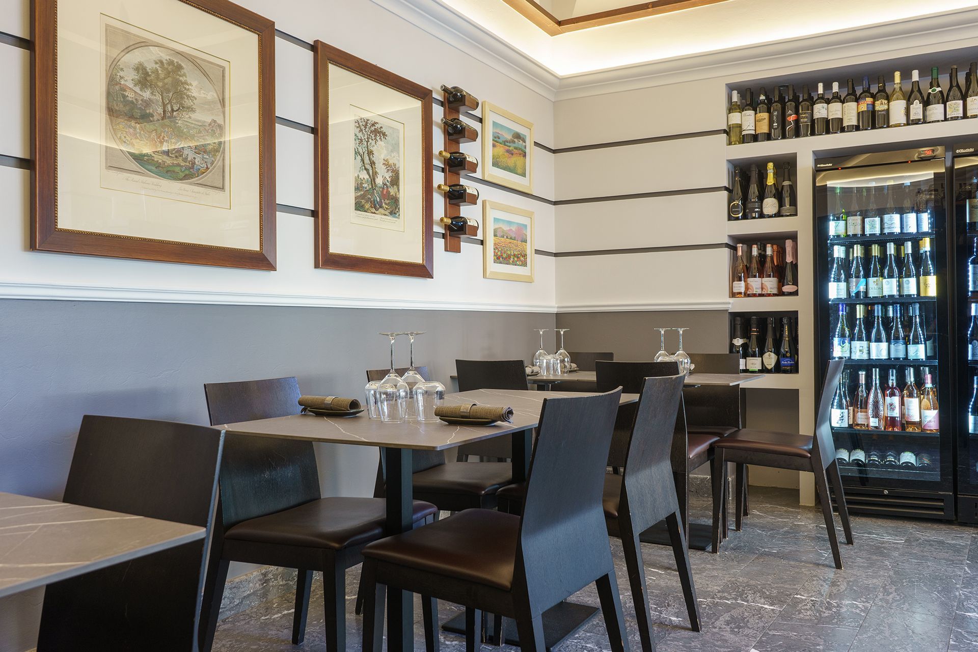 Piccolo ristorante moderno con tavoli scuri, opere d'arte alle pareti, scaffali per il vino e un refrigeratore per bevande in vetro.