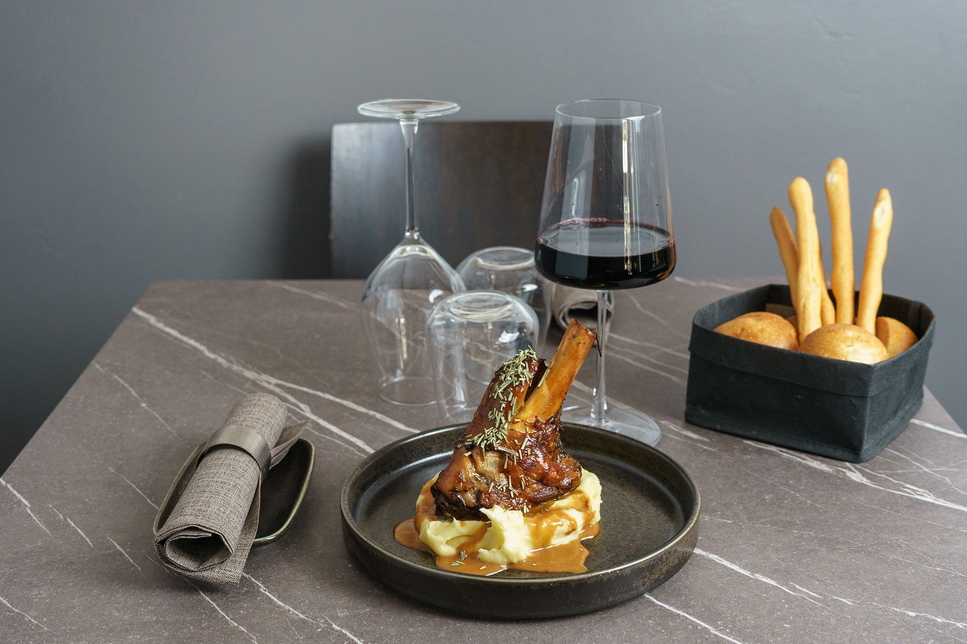 Elegante dessert servito al piatto con salsa scura, calice da vino, tovagliolo e grissini su un tavolo grigio.