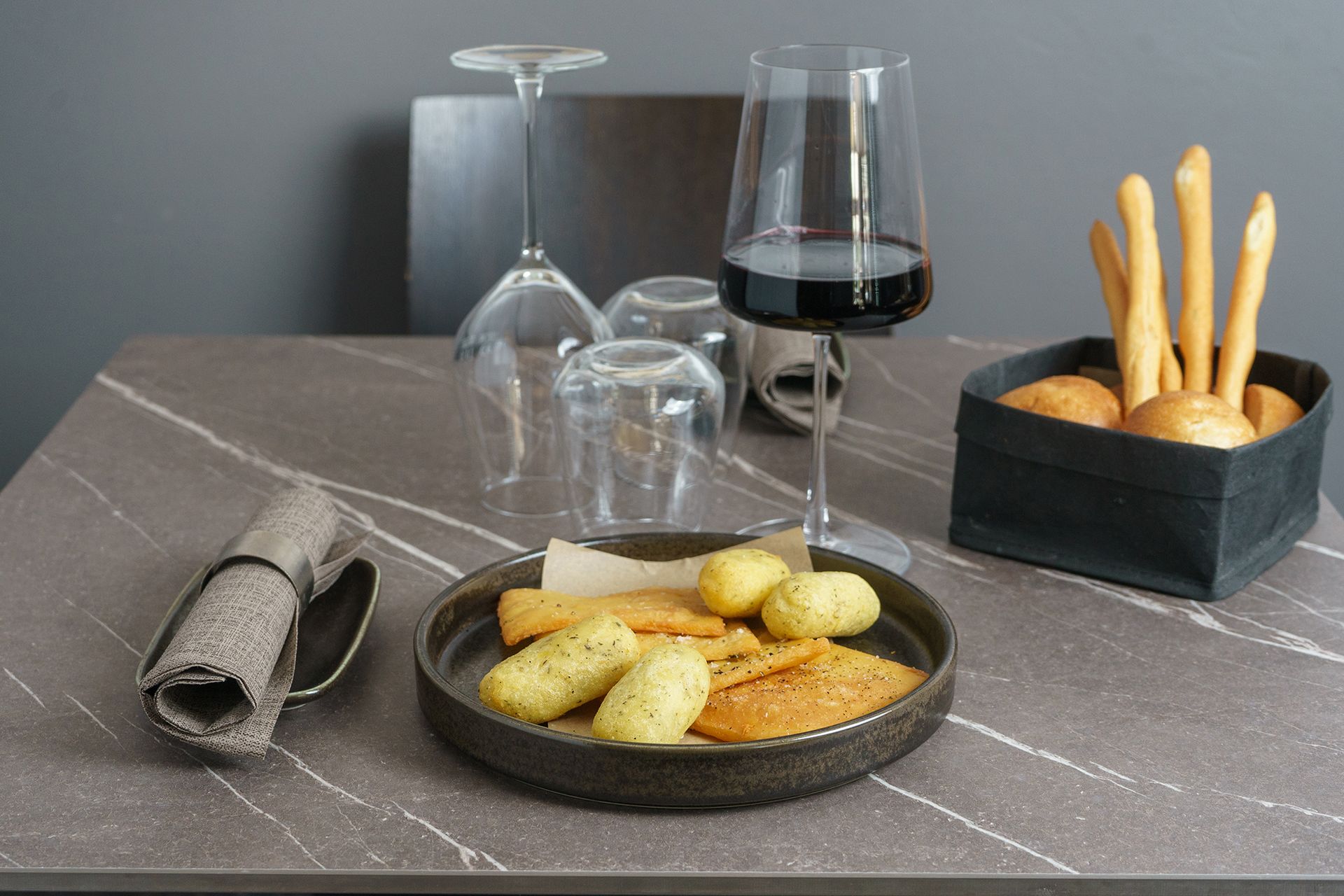 Una tavola apparecchiata con pane, cracker, vino, bicchieri capovolti e un cestino del pane su una superficie grigia.