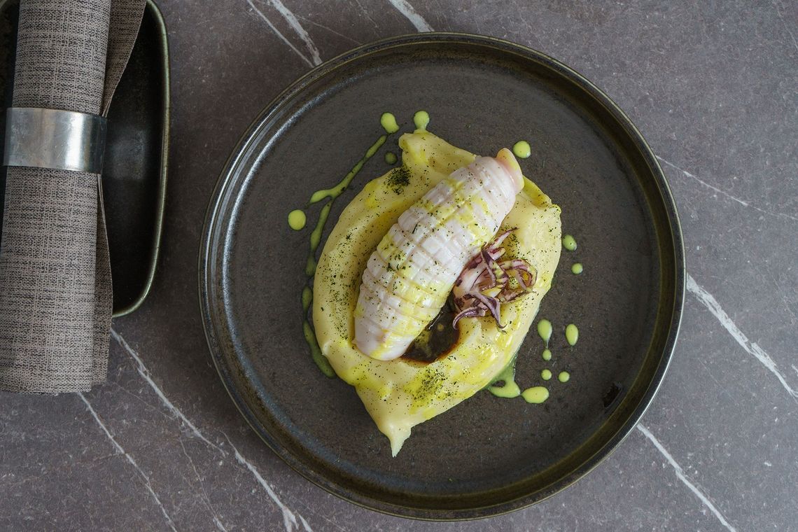 Piatto gourmet impiattato con pesce a fette in salsa verde, accanto a un coltello su un tavolo di pietra grigia.