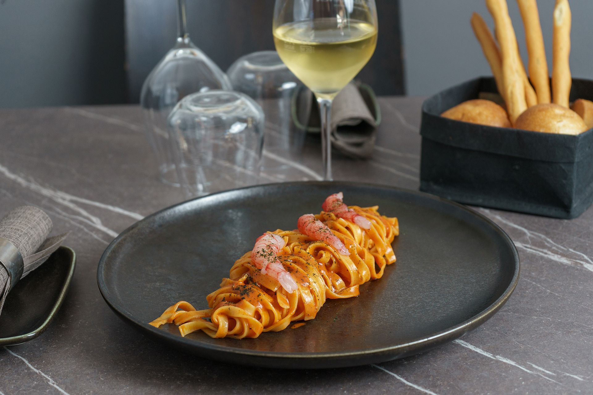 Pasta su un piatto nero con bicchieri da vino e grissini su un tavolo di marmo scuro.