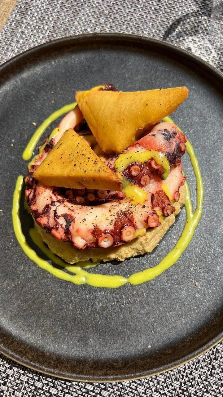 Polpo servito su un piatto di ceramica scura con triangolini croccanti e salsa verde a spirale.