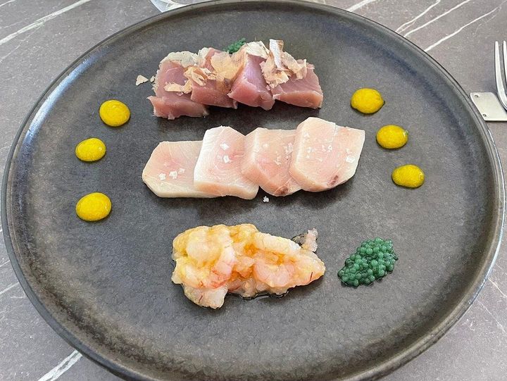 Assortimento di pezzi di sushi sashimi disposti su un piatto scuro con gocce di salsa gialla e guarnizione verde.