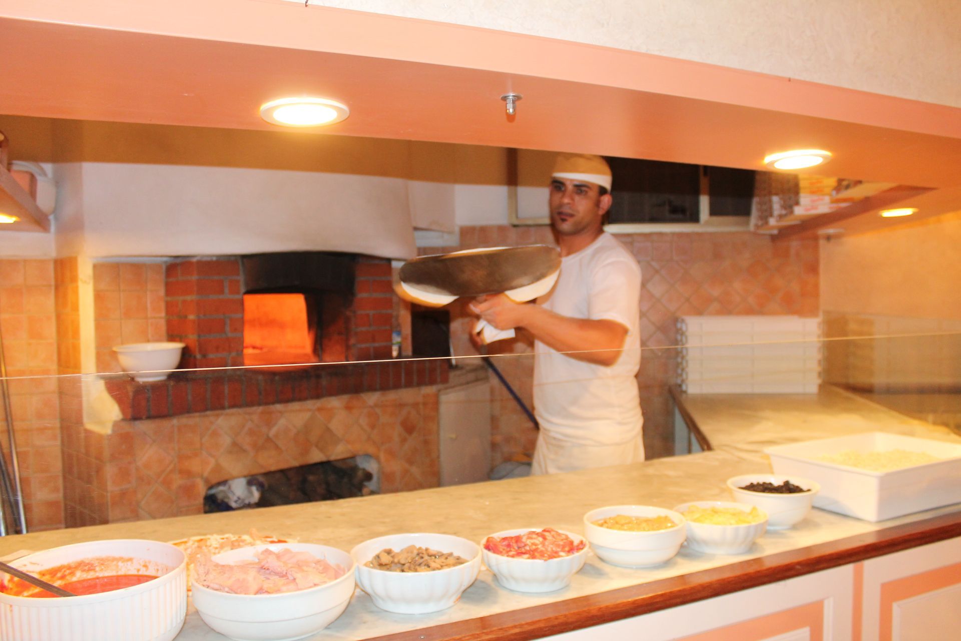 Una persona tiene in mano una pizza vicino a un forno a legna nella cucina di un ristorante, con i condimenti sul bancone.