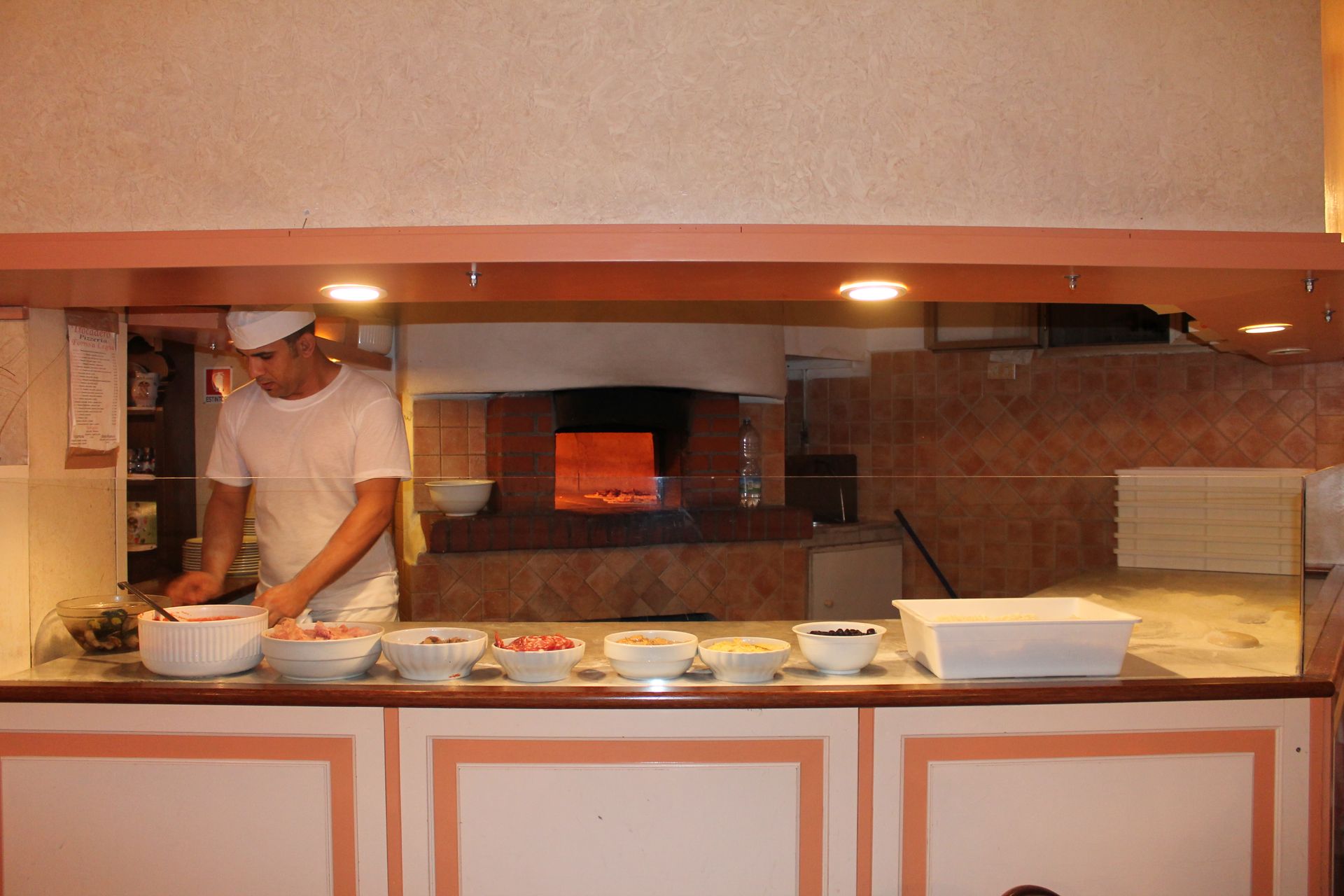 Chef che prepara la pizza al bancone, ingredienti nelle ciotole, forno a legna sullo sfondo.