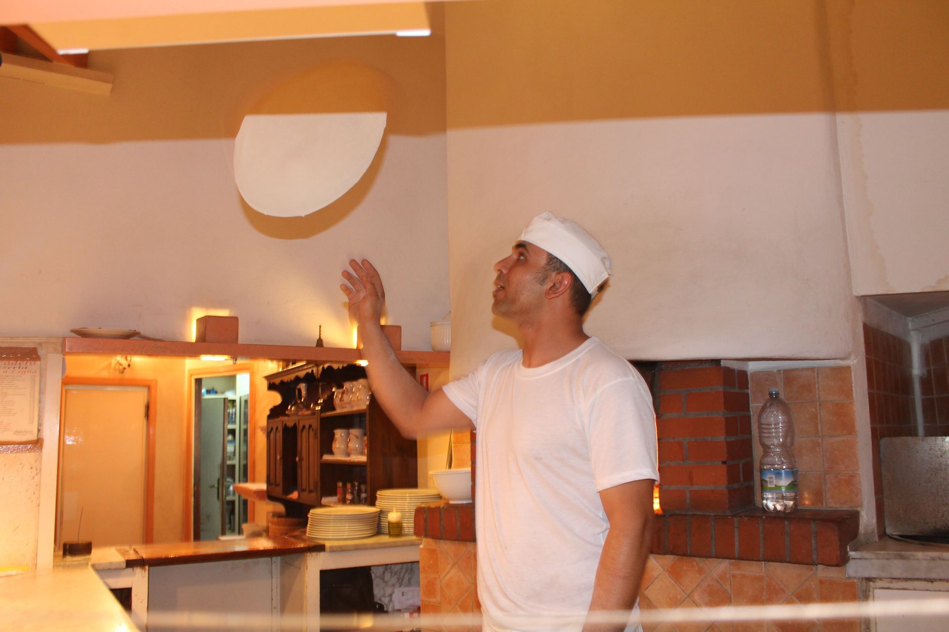 Uno chef in uniforme bianca lancia l'impasto della pizza in un locale chiuso, vicino a un forno a legna.