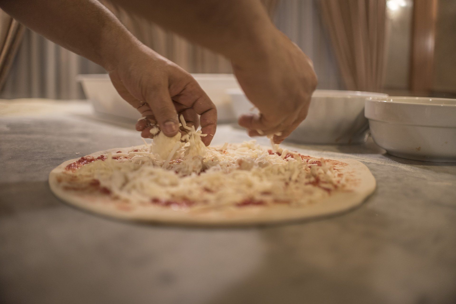 Mani che cospargono di formaggio una base per pizza con salsa rossa, accanto a ciotole bianche su un tavolo.