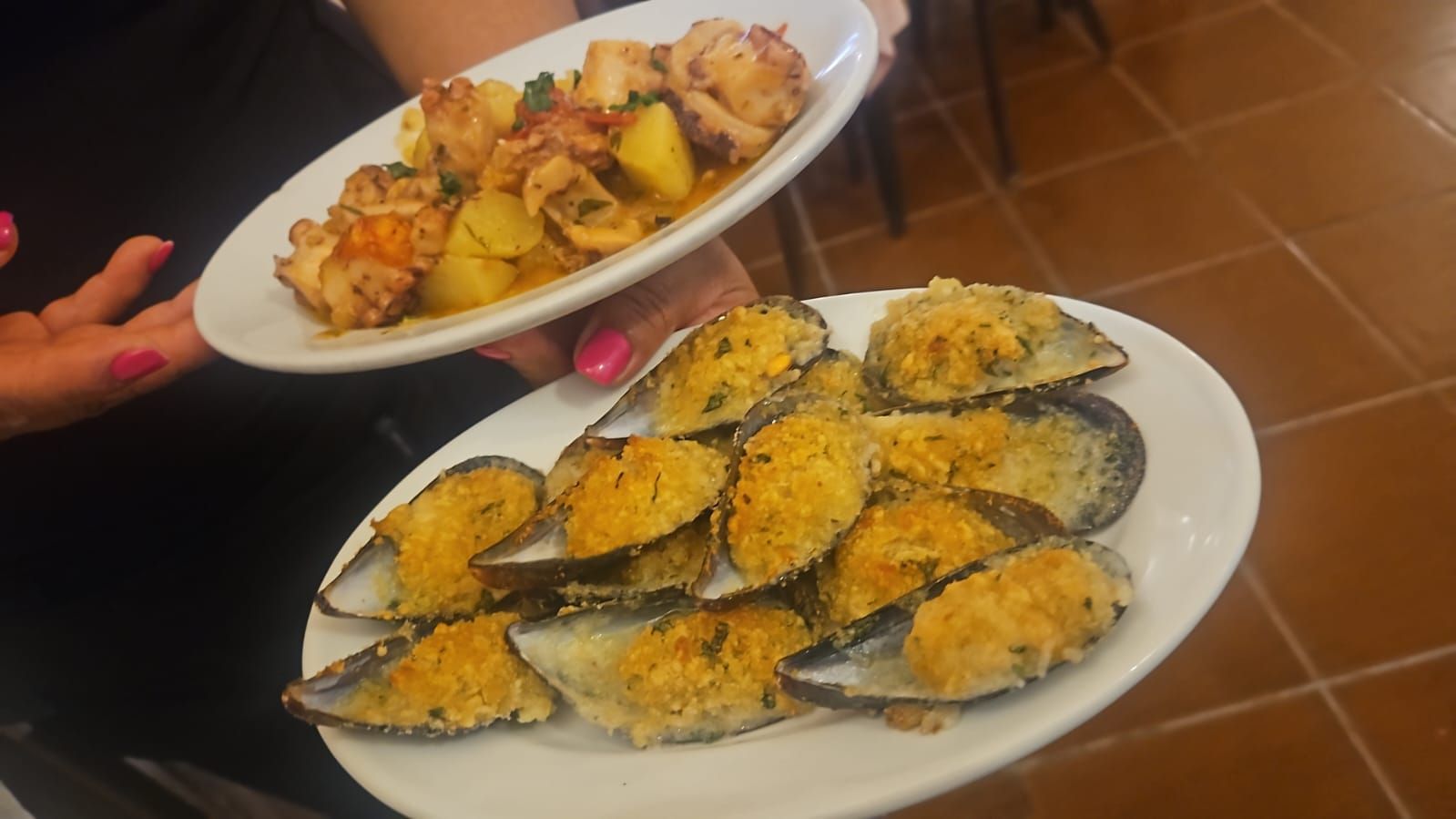 Donna che tiene in mano due piatti di cibo: cozze al forno e un piatto di frutti di mare con patate.