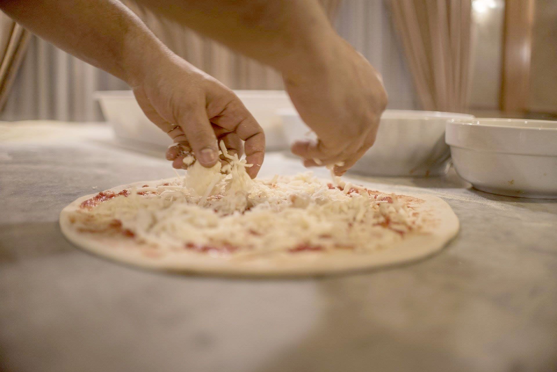 Chef che prepara la pizza al bancone, ingredienti nelle ciotole, forno a legna sullo sfondo.