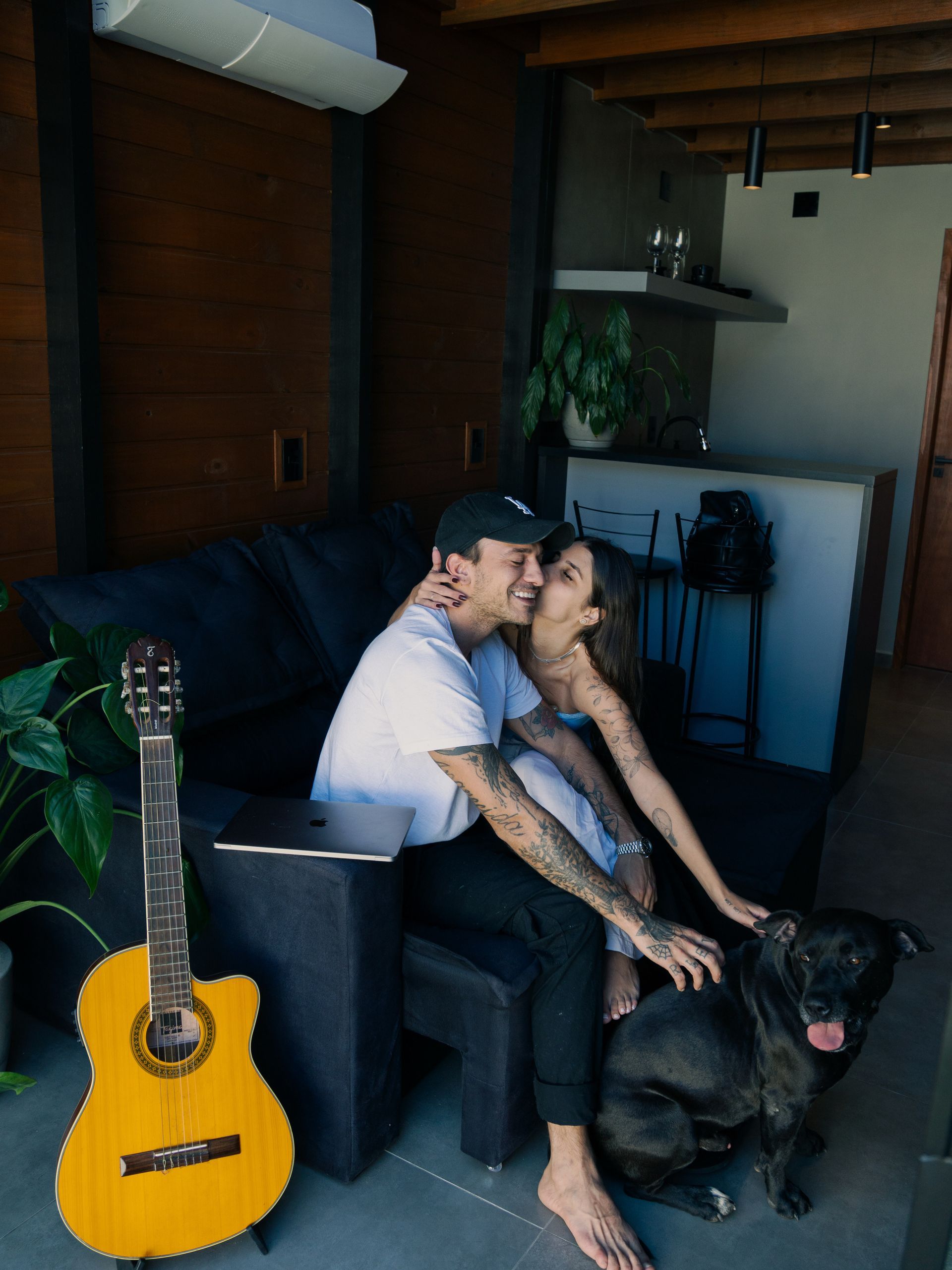 Casal sentado em um sofá com um cachorro preto e um violão em uma sala aconchegante.