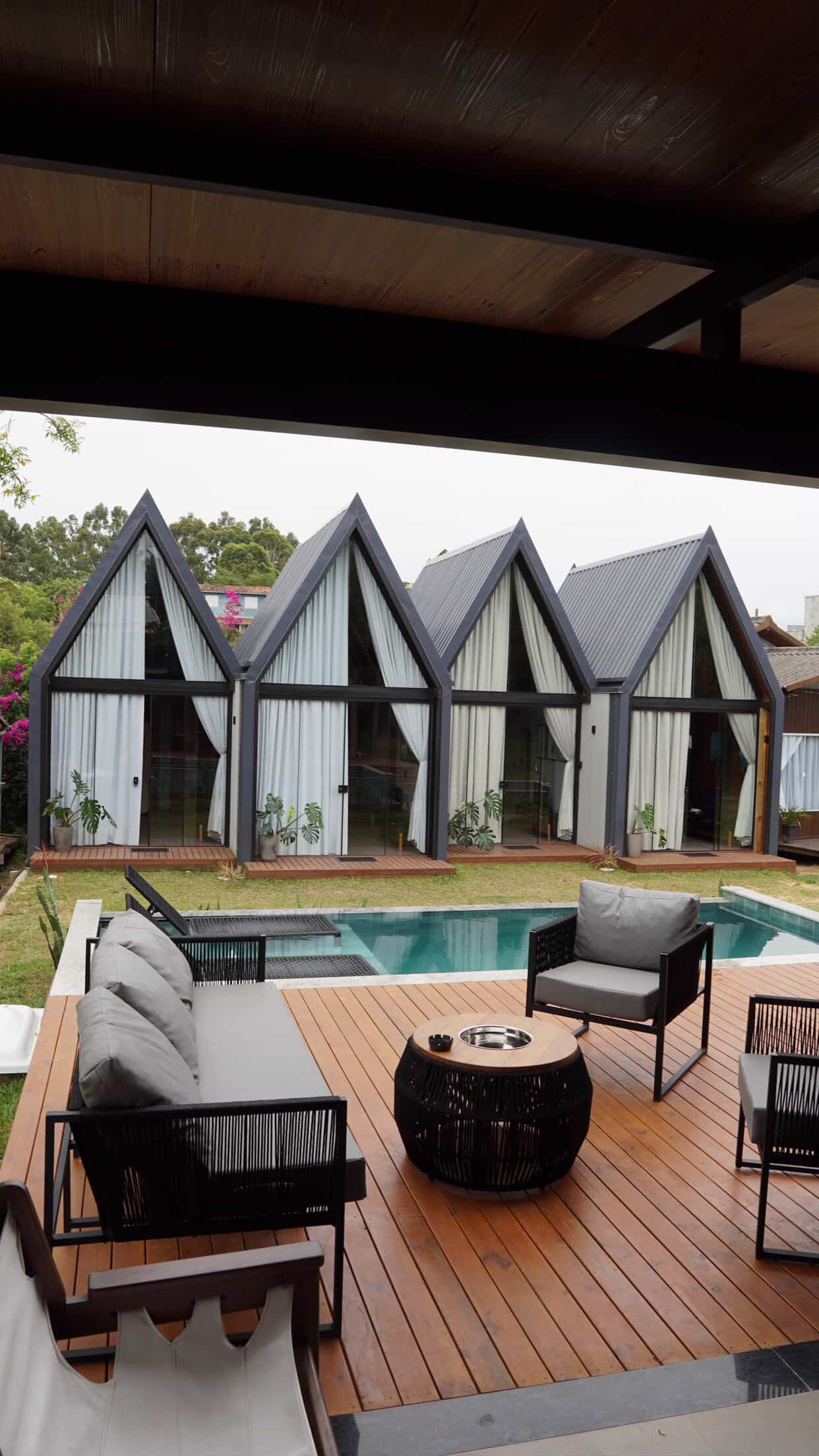 Pátio moderno com piscina, espreguiçadeiras pretas, lareira externa e chalés com telhado triangular nos fundos.