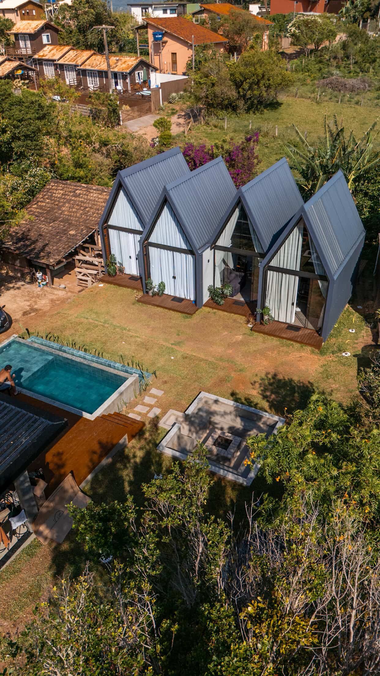 Vista aérea de uma pequena casa de campo branca com telhado azul ao lado de uma piscina turquesa e vegetação exuberante.