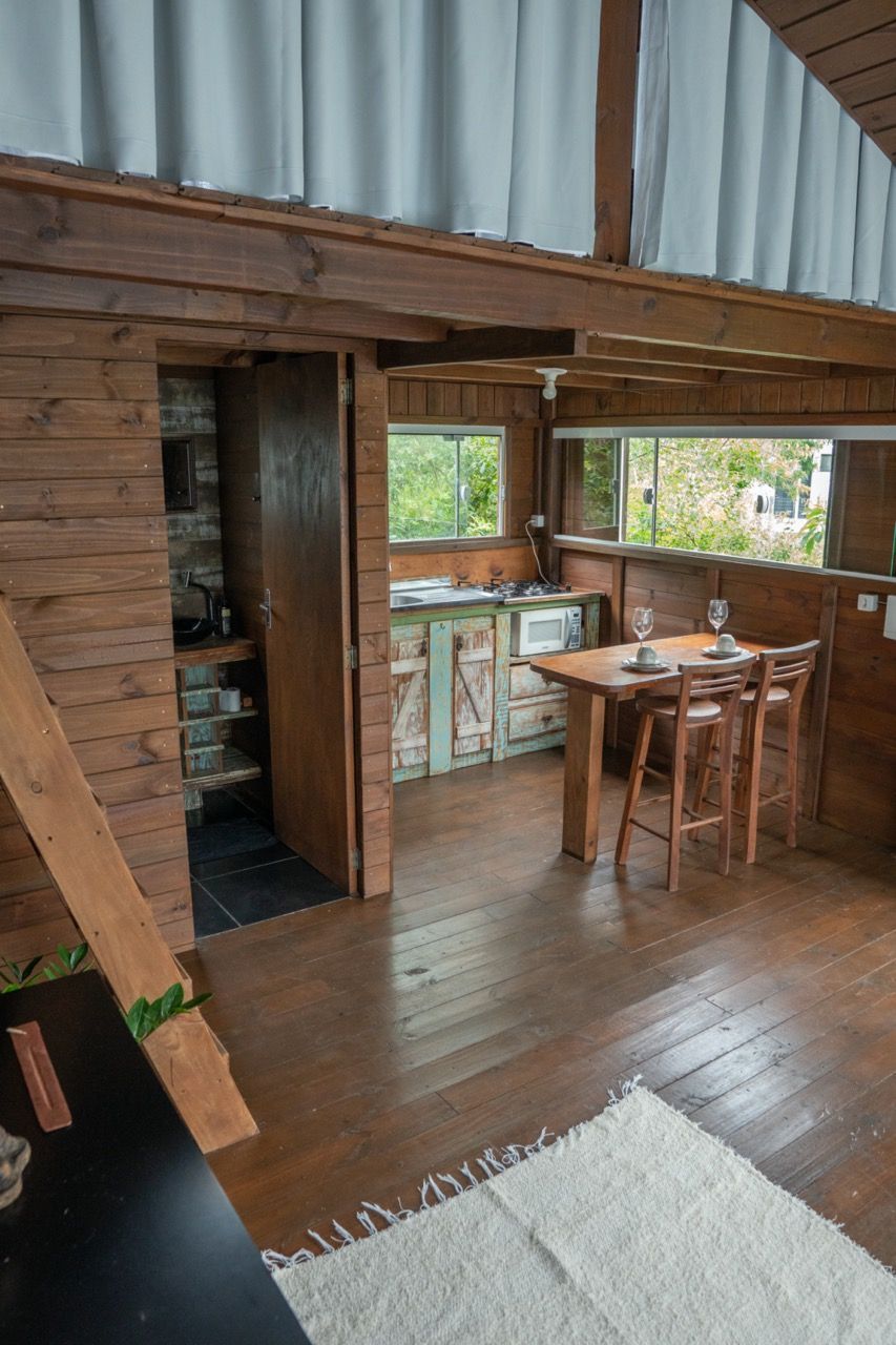 Interior rústico de loft em madeira com uma pequena mesa, cadeiras e grandes janelas com vista para a vegetação.