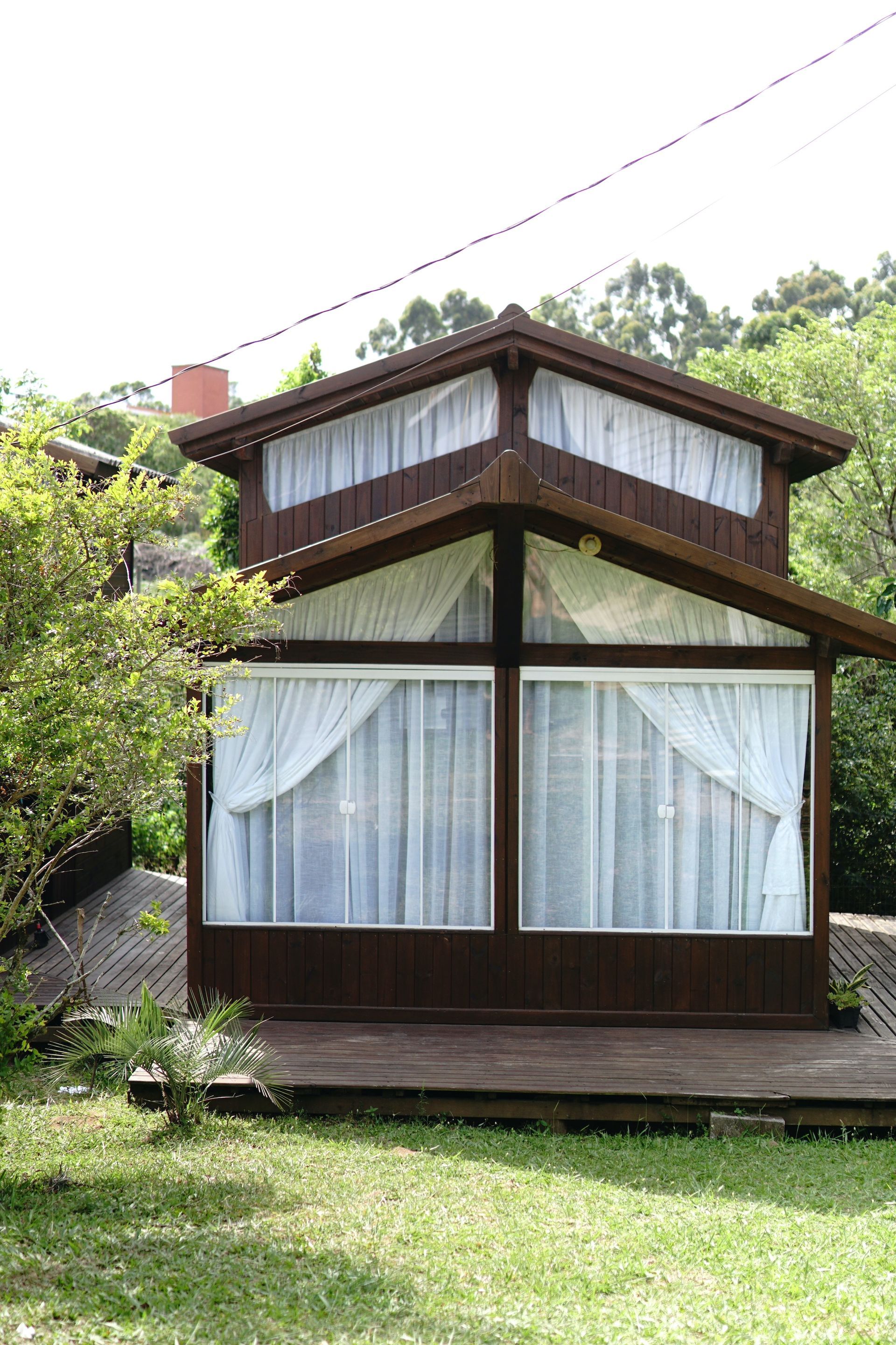 Pequena cabana de madeira com grandes janelas frontais, cortinas brancas e um quintal gramado rodeado por árvores.