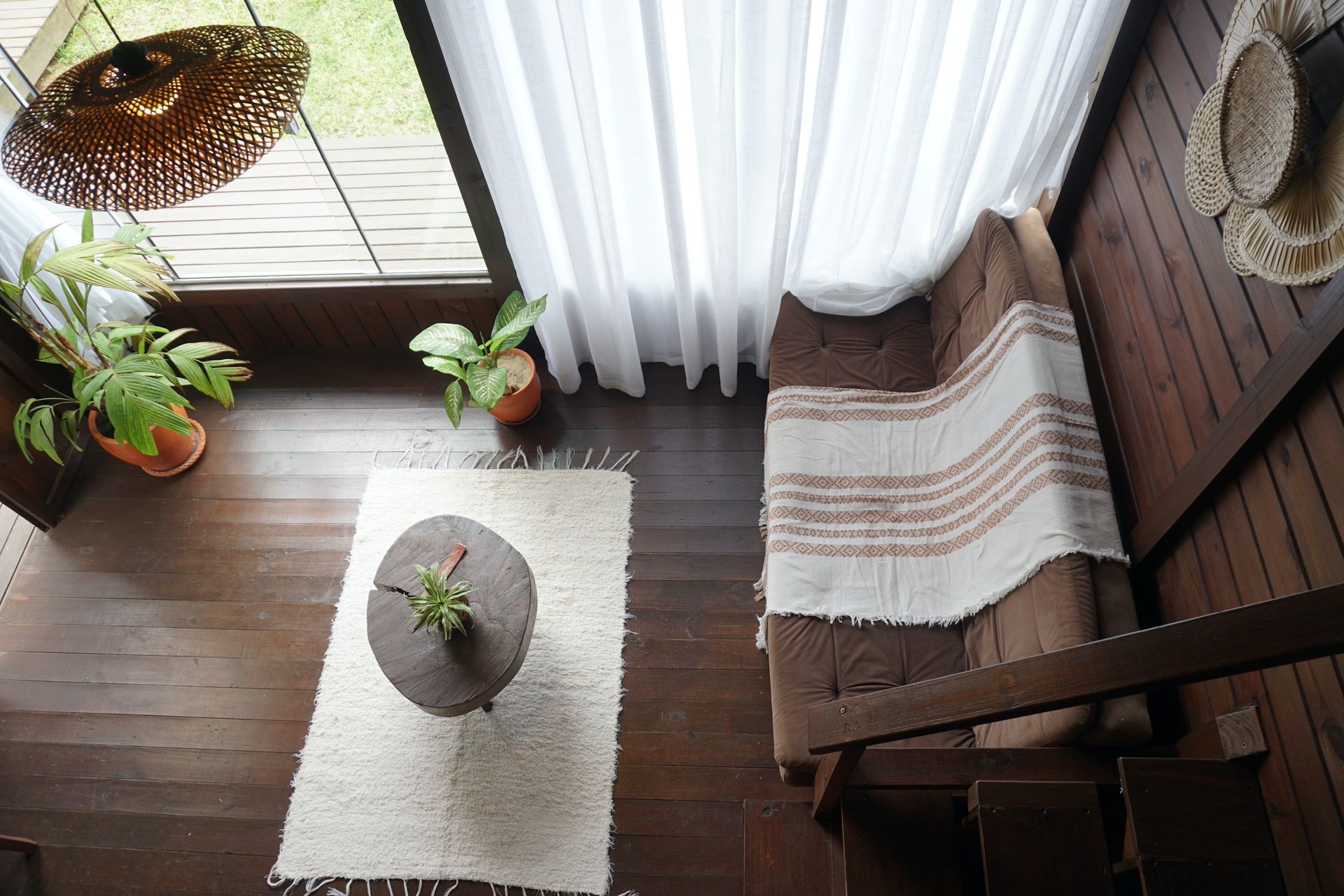 Cantinho aconchegante da sala de estar com cadeira de vime, cortinas brancas, tapete e vasos de plantas perto de uma janela ensolarada.