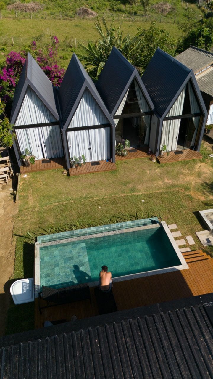 Vista aérea de pequenas cabanas em forma de A ao lado de uma piscina turquesa, com uma pessoa na água e um gramado.
