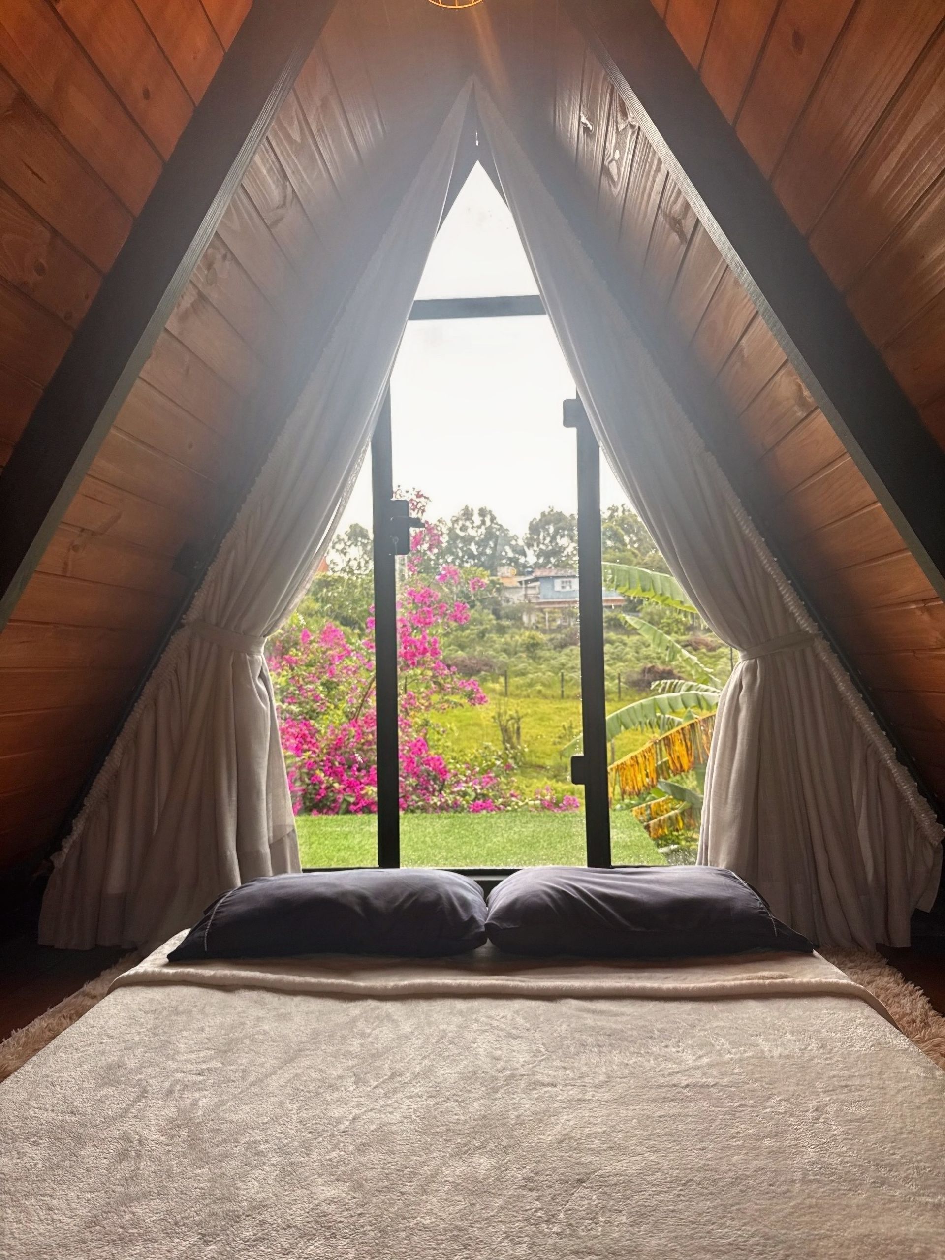 Quarto em cabana em forma de A com cortinas abertas que emolduram um jardim e flores coloridas do lado de fora.